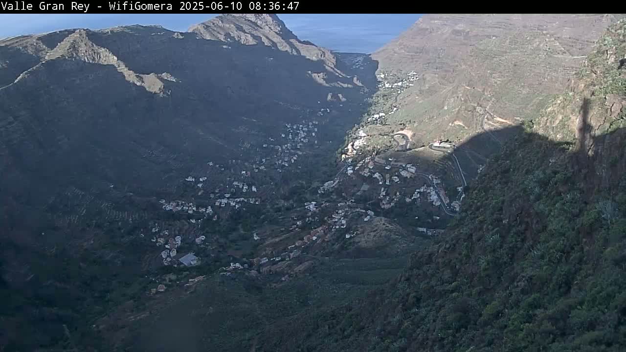 A high-angle view shows a mountainous valley town nestled between steep, dark-green slopes on a sunny day.