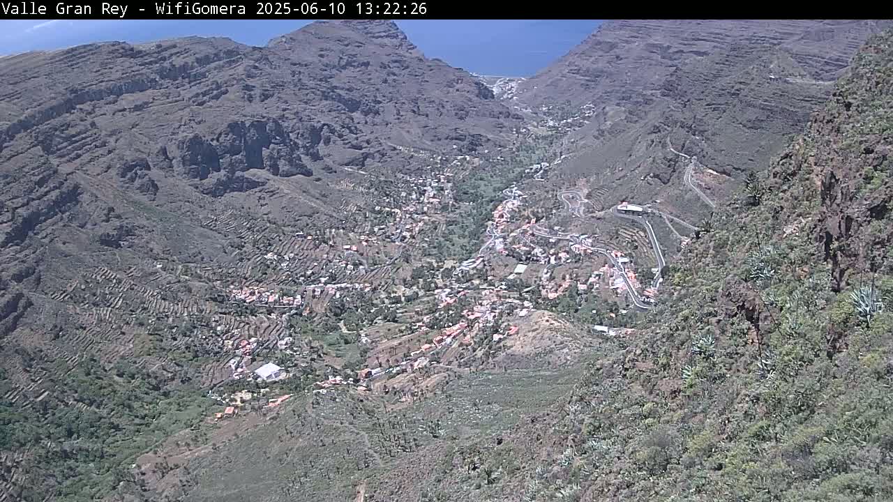 A high-angle view on a sunny day shows a small town nestled in a steep, rocky valley with terraced hillsides and winding roads.