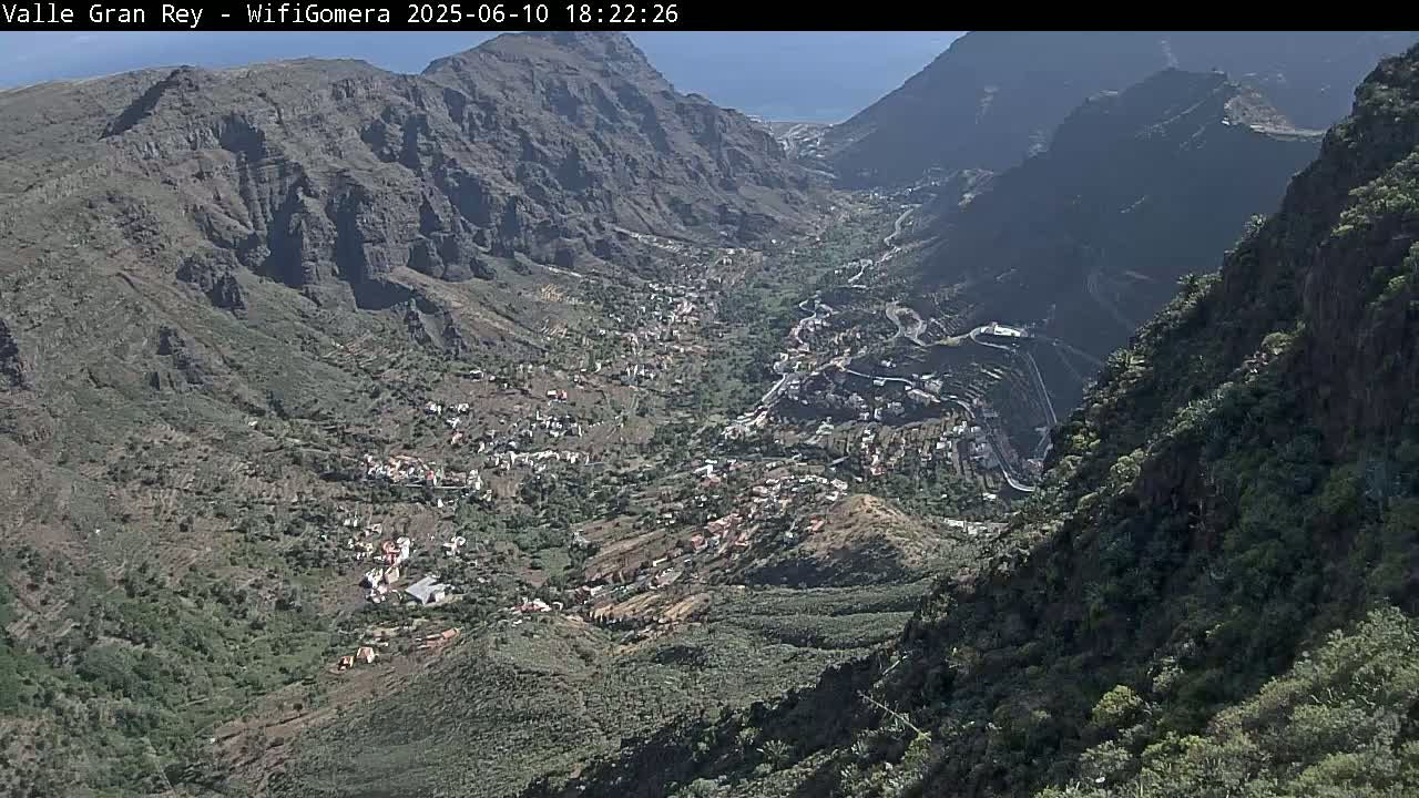 A high-angle view shows a valley nestled between steep, rocky mountains, with a small town situated in the valley floor on a sunny day.