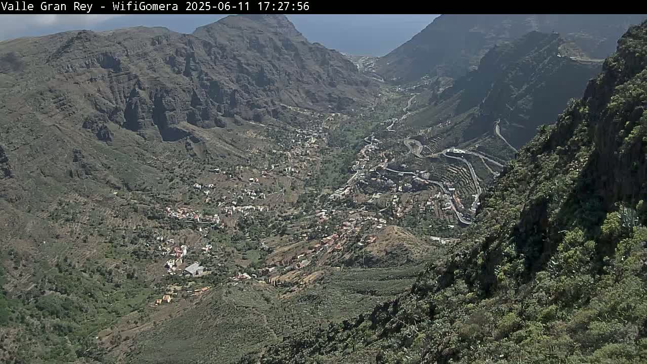 A high-angle view shows a valley town nestled between steep, rocky mountains on a sunny day.