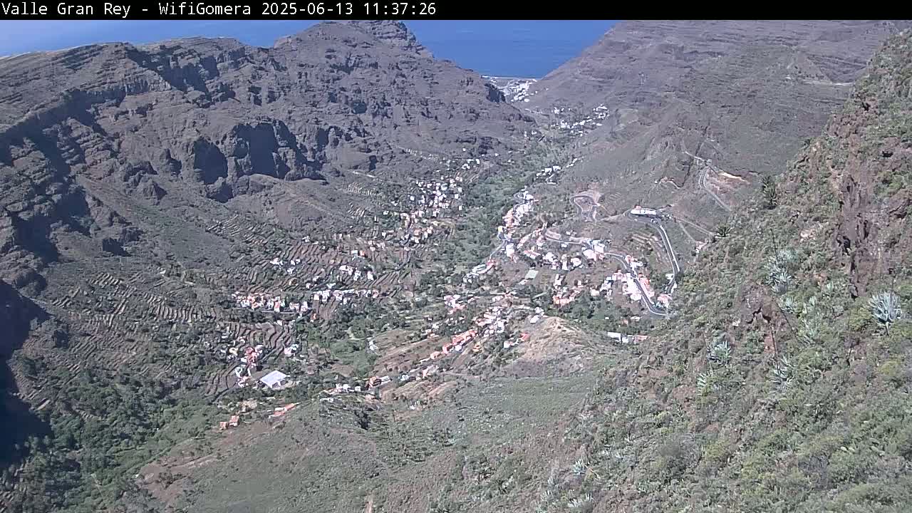 A high-angle view shows a small town nestled in a mountainous valley on a sunny day, with the ocean visible in the distance.