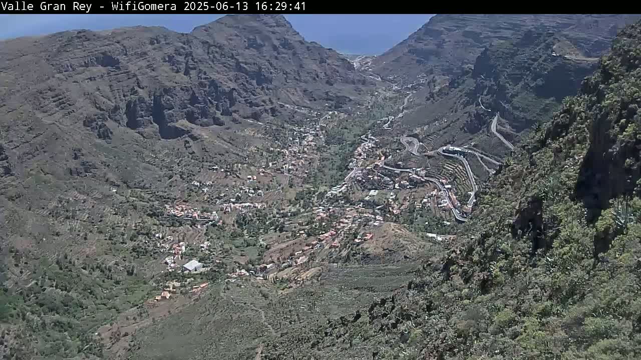 A high-angle view on a sunny day shows a mountainous valley with a small town nestled within, winding roads snaking through the landscape.