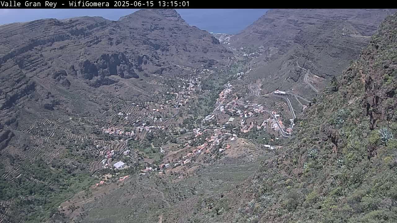A high-angle view on a sunny day shows a small town nestled in a mountainous valley, with terraced farmland surrounding it.