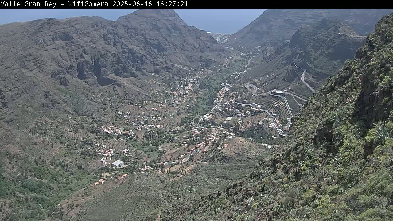 A high-angle view shows a mountainous valley town with winding roads on a sunny day, overlooking a village nestled amongst terraced hillsides.