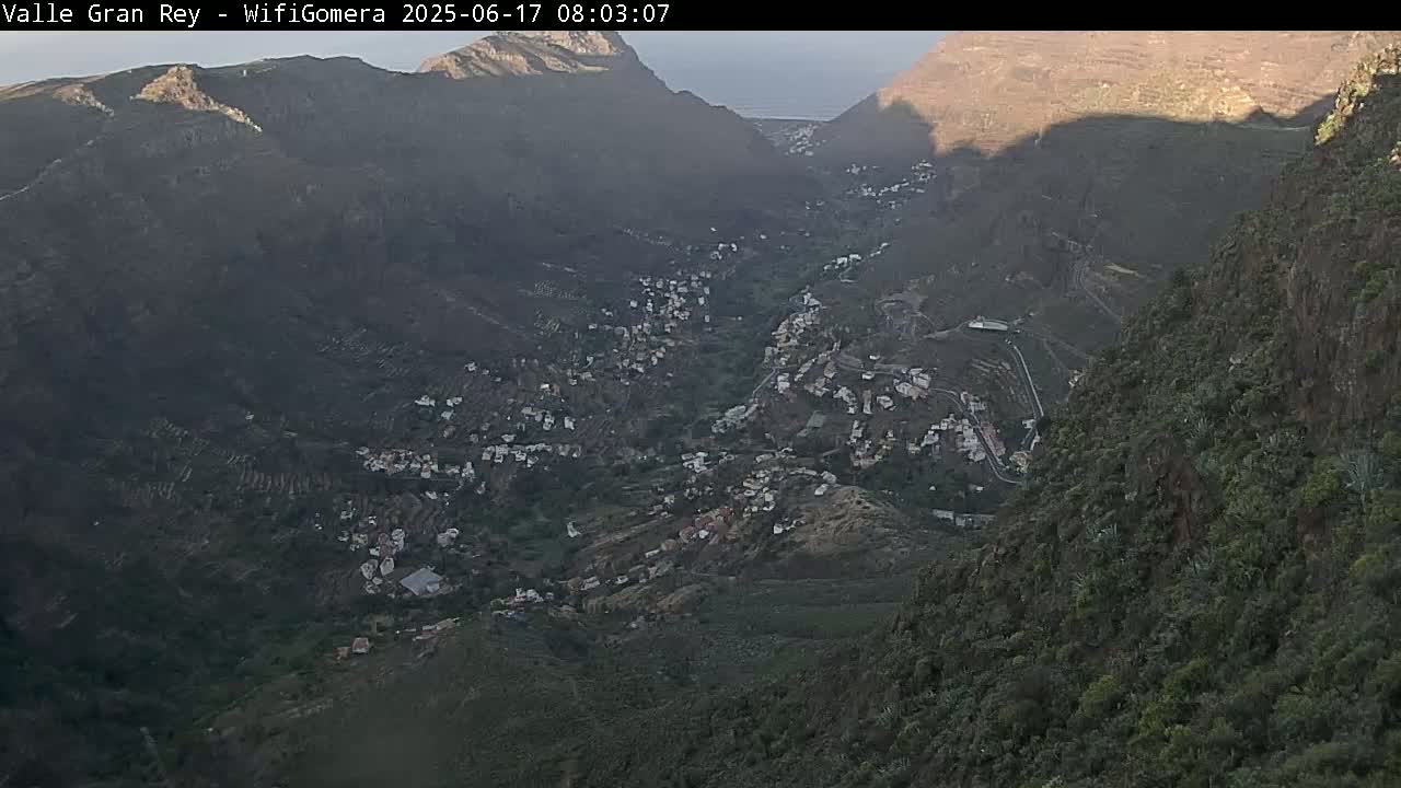 A mountainous valley town is visible at dawn, nestled between steep, dark green and brown hillsides.