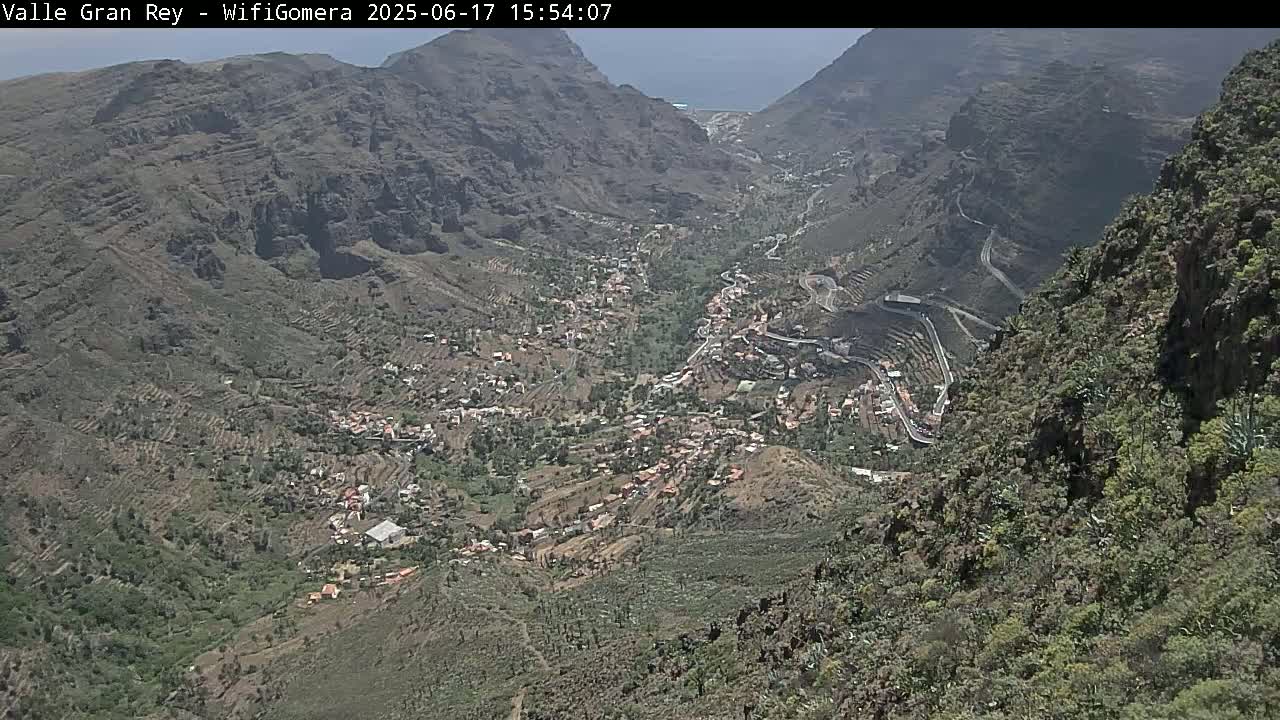 An aerial view shows a valley town nestled between steep, rocky mountainsides on a sunny day.