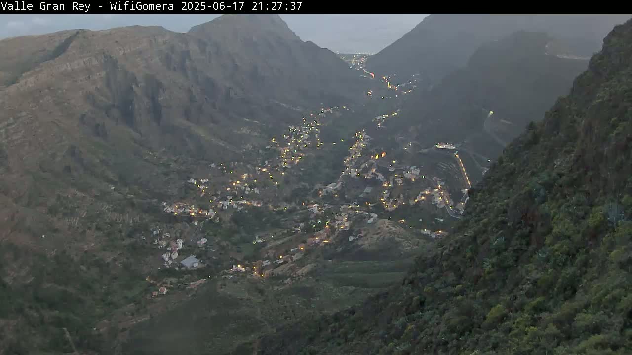 A valley town nestled between dark, mountainous slopes is illuminated at night under a dusky sky.