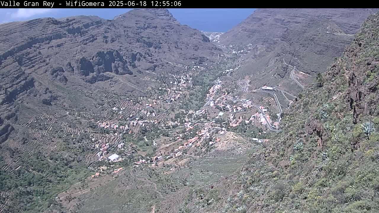 A high-angle view shows a small town nestled in a steep, rocky valley under a clear blue sky.