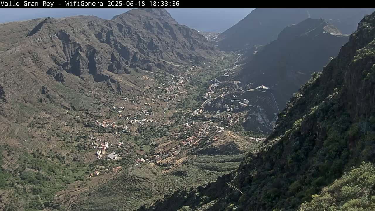 A high-angle view shows a valley nestled between steep, rocky mountains under a sunny sky, with a small town situated in the valley bottom.