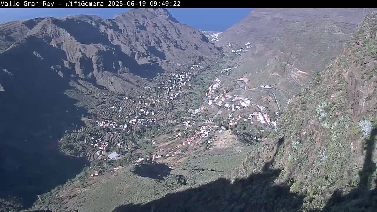 A high-angle view shows a sunlit valley nestled between steep, rocky mountains, with a village situated in the valley below.