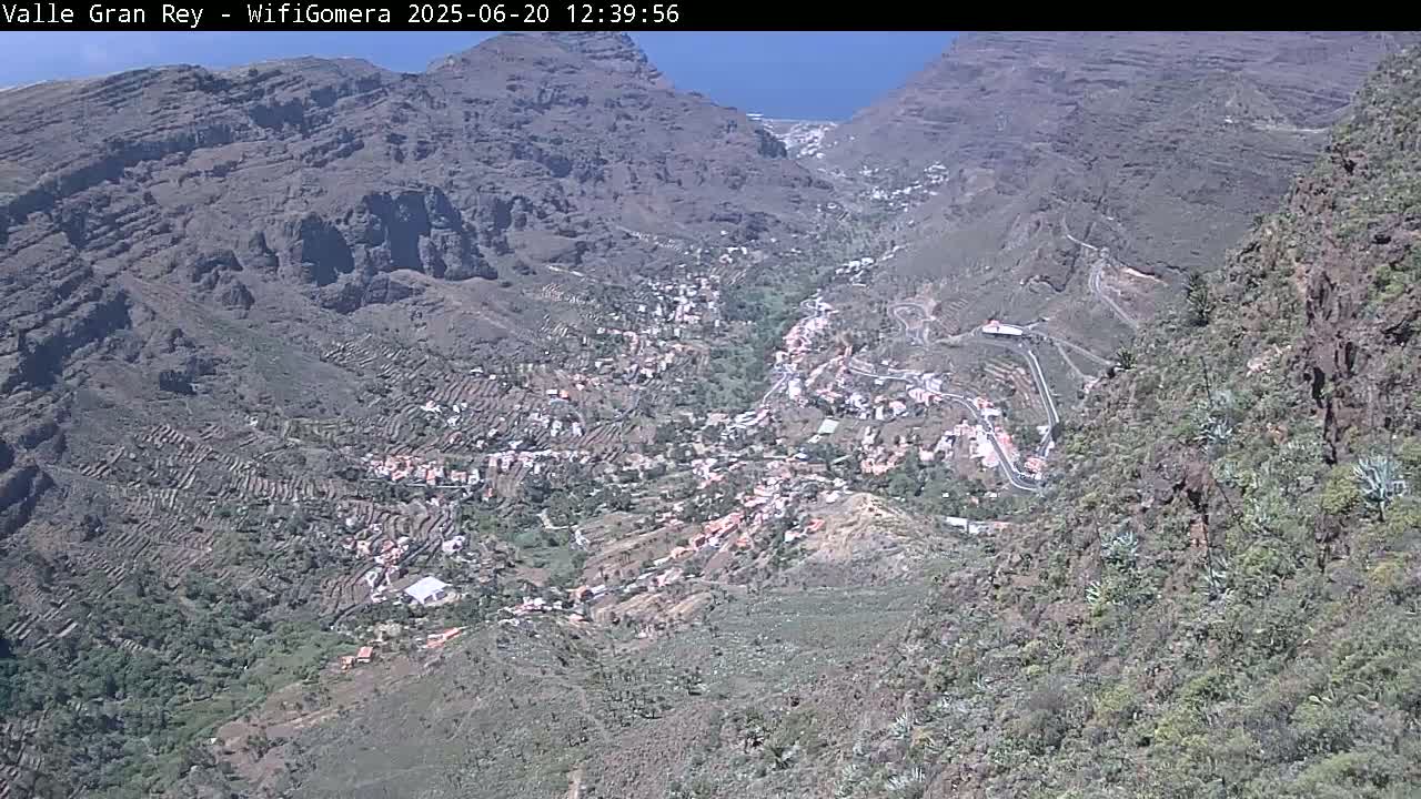 A high-angle view shows a village nestled in a mountainous valley on a sunny day, with terraced farmland visible on the slopes.