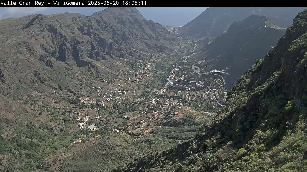 A high-angle view shows a valley town nestled between steep, rocky mountains on a sunny day.