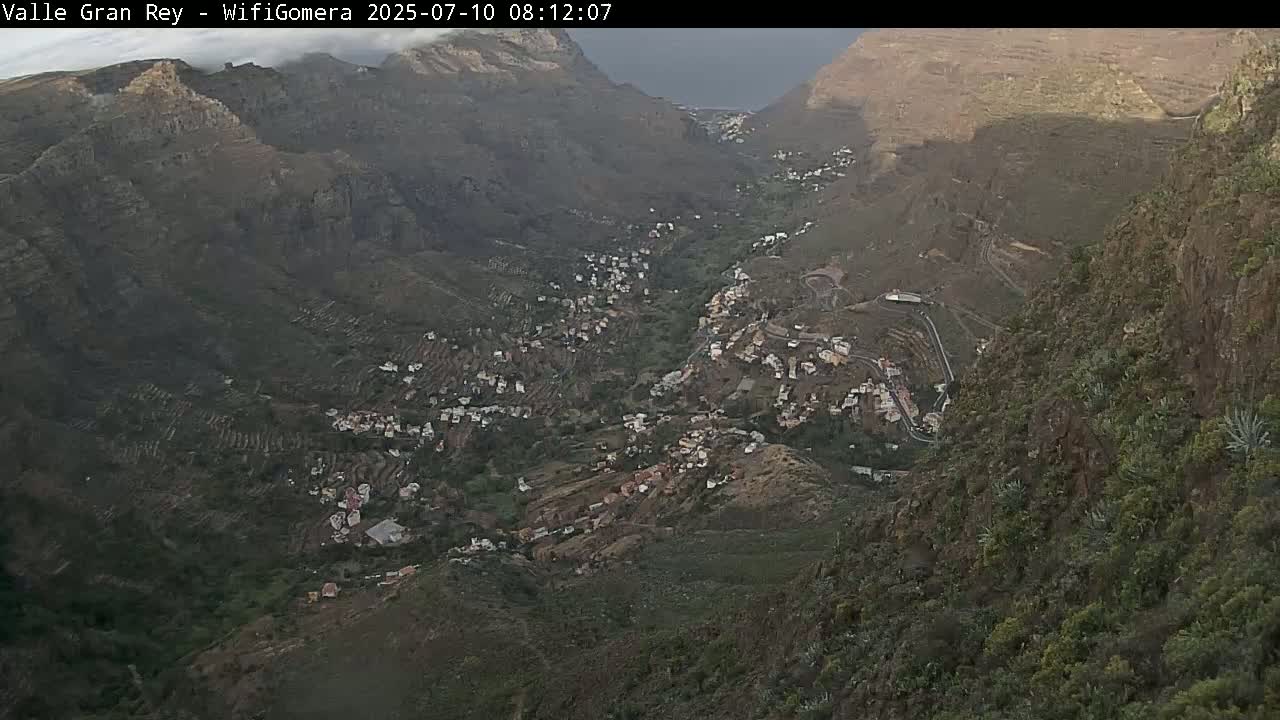 A high-angle view shows a village nestled in a steep, mountainous valley under a partly cloudy sky.