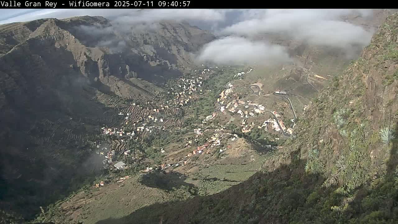A partly cloudy day reveals a mountainous valley town with terraced farmland, winding roads, and cliffsides dotted with vegetation.