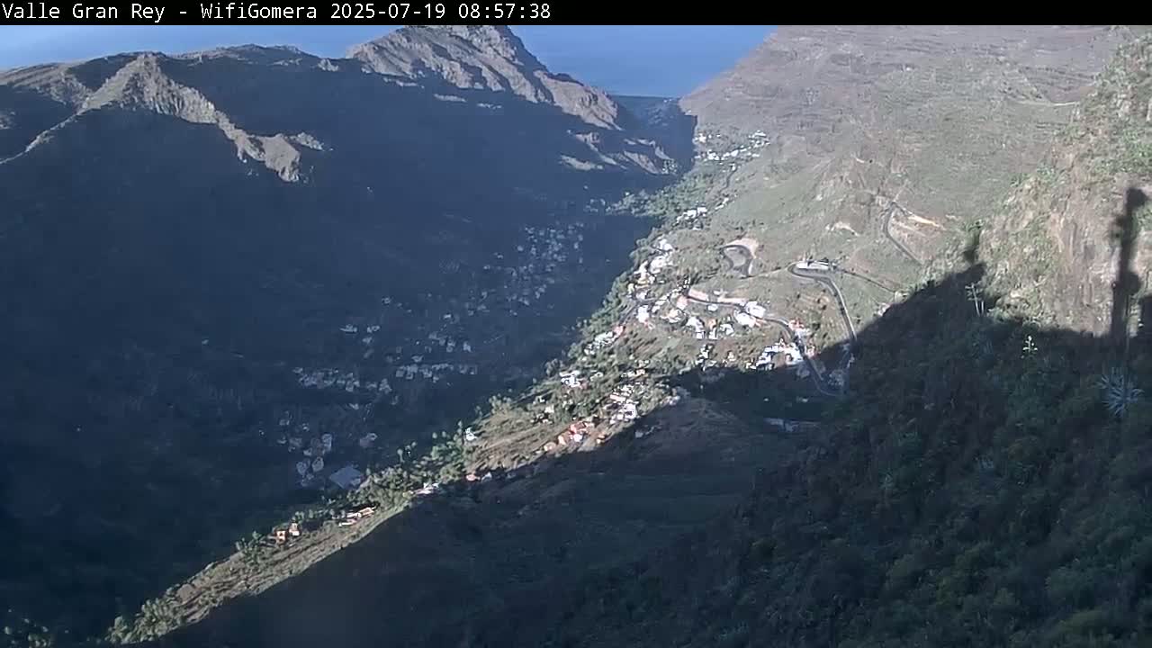 A high-angle view shows a sunlit valley town nestled between steep, shadowed mountainsides under a mostly clear sky.