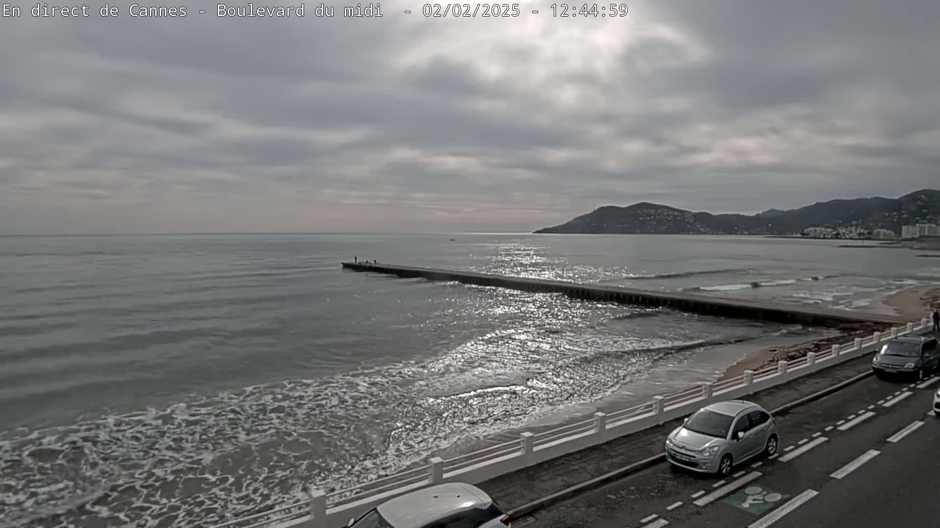 A mostly cloudy day shows a long pier extending into a gently waving sea, with cars parked along a coastal road and distant hills under a gray sky.