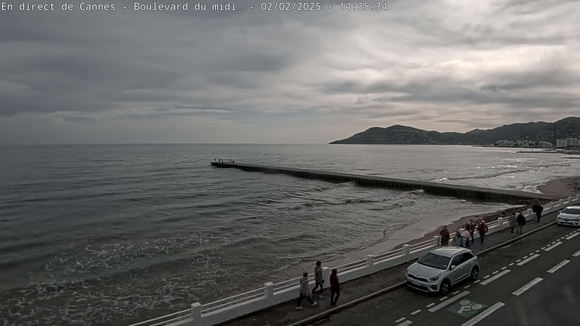 A long pier extends into a gray, choppy sea under a cloudy sky, with people walking along a shoreline road next to a beach.