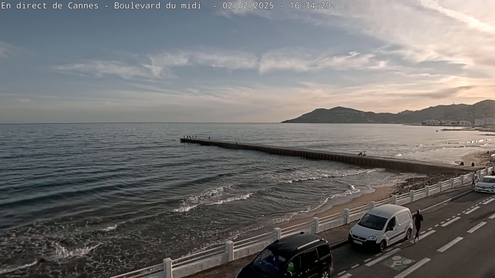 A long pier extends into a calm sea under a partly cloudy sky, with a sandy beach and a road with cars visible in the foreground.