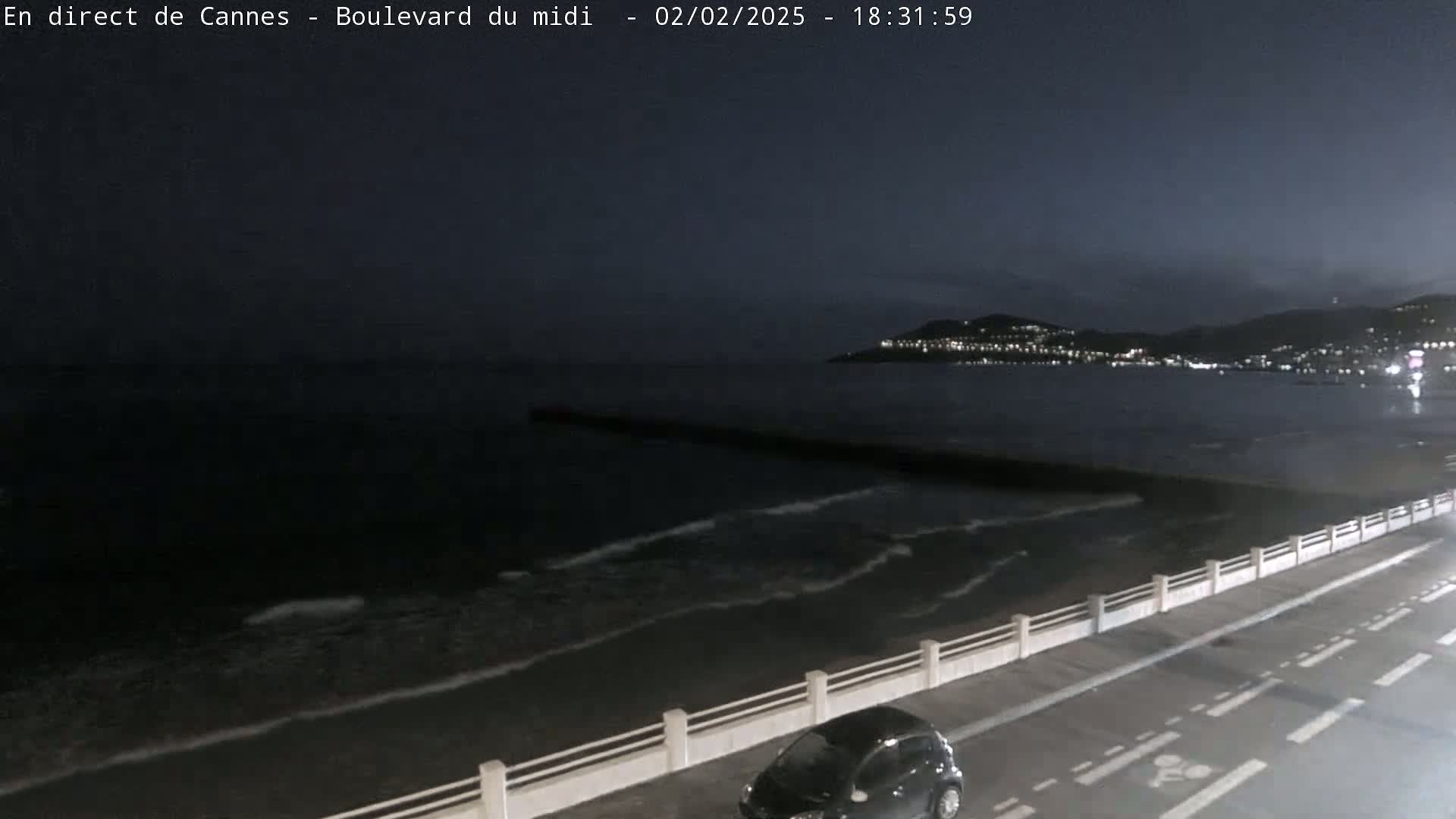 A nighttime coastal scene shows dark, gently rolling waves lapping a shoreline with a roadway and a single car, and a city's lights visible in the distance.