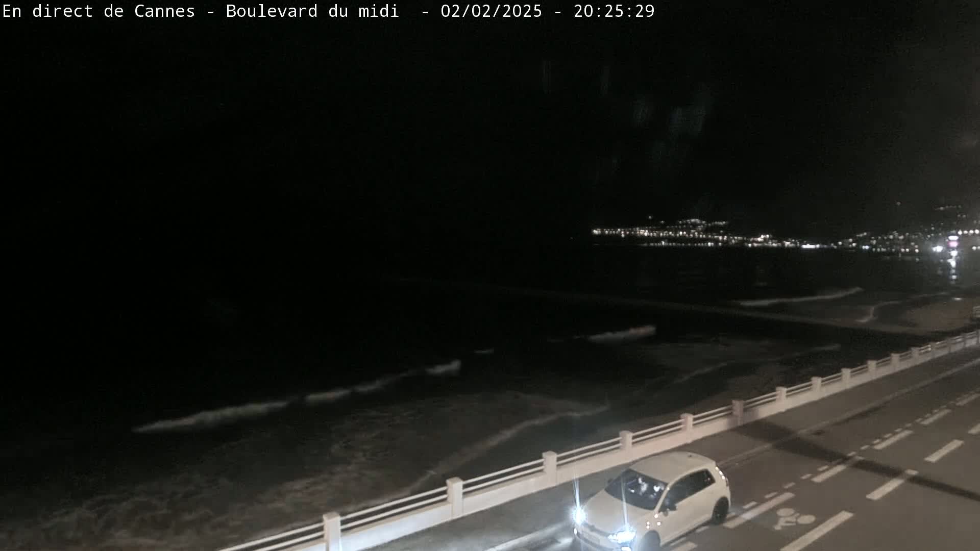A nighttime view of a dark ocean with small waves gently lapping the shore, a road with a single white car, and a distant city skyline illuminated with lights.