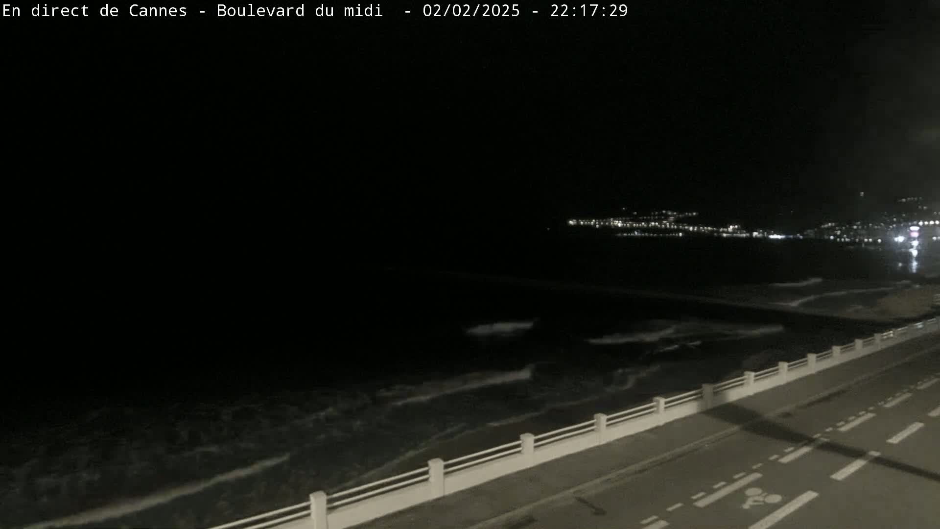 A nighttime view shows dark ocean waves lapping against a seawall next to a roadway.