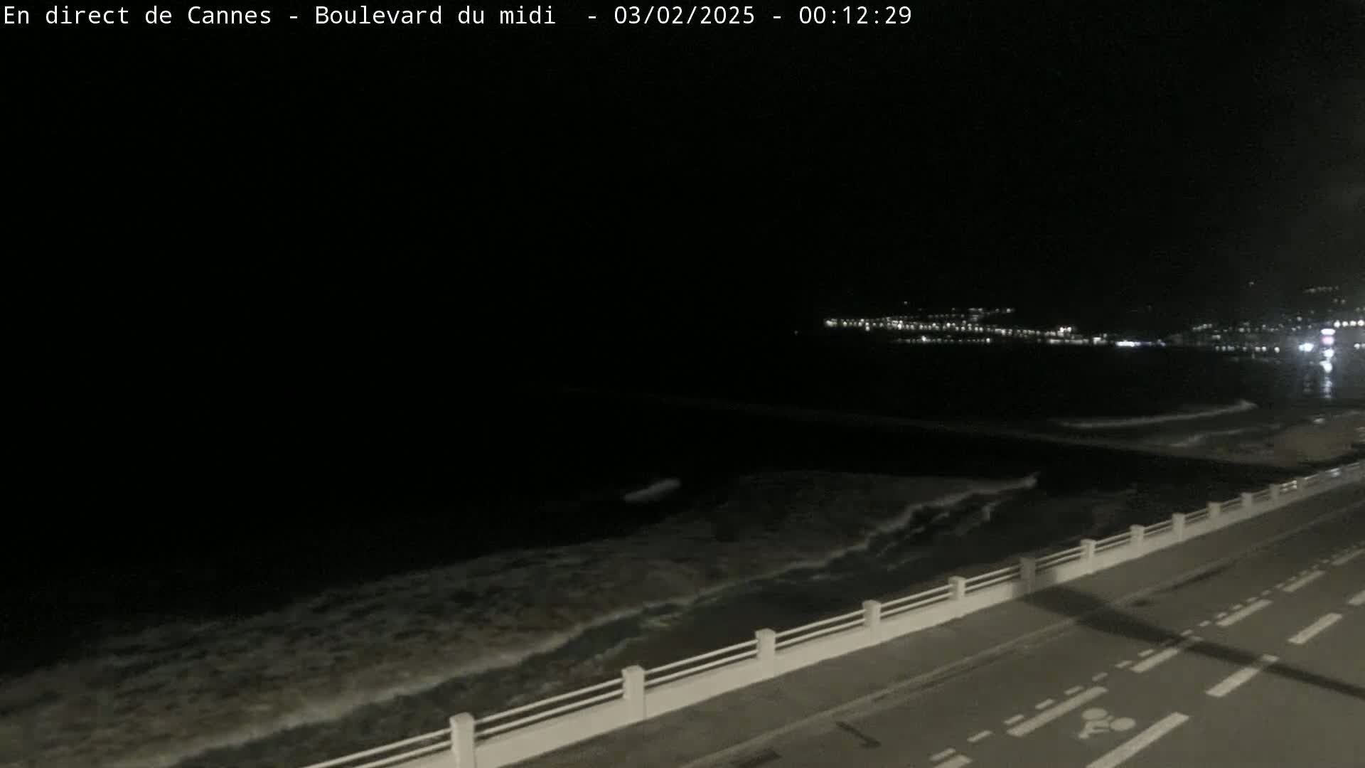 A nighttime coastal scene shows dark water with small waves lapping against a seawall next to a road.