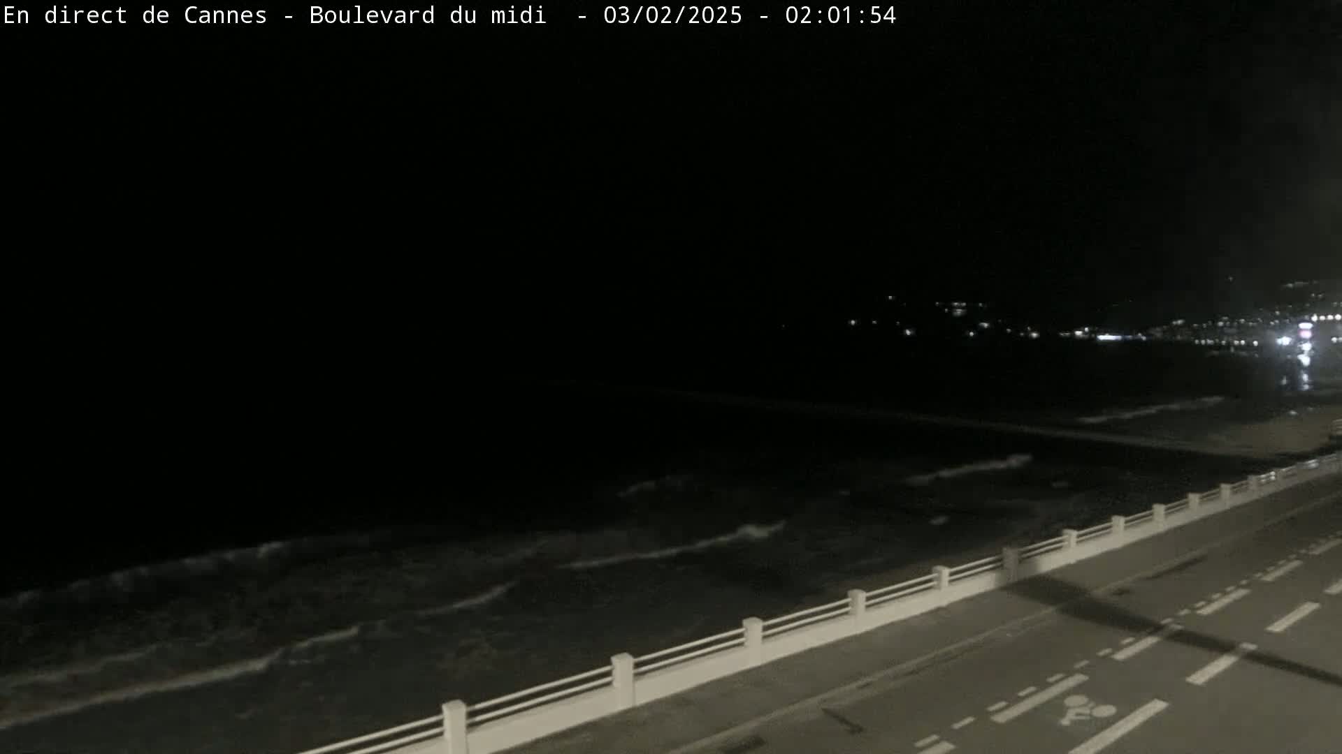 A dark nighttime scene shows a paved road next to a low white railing overlooking a gently lapping ocean under a clear, dark sky.