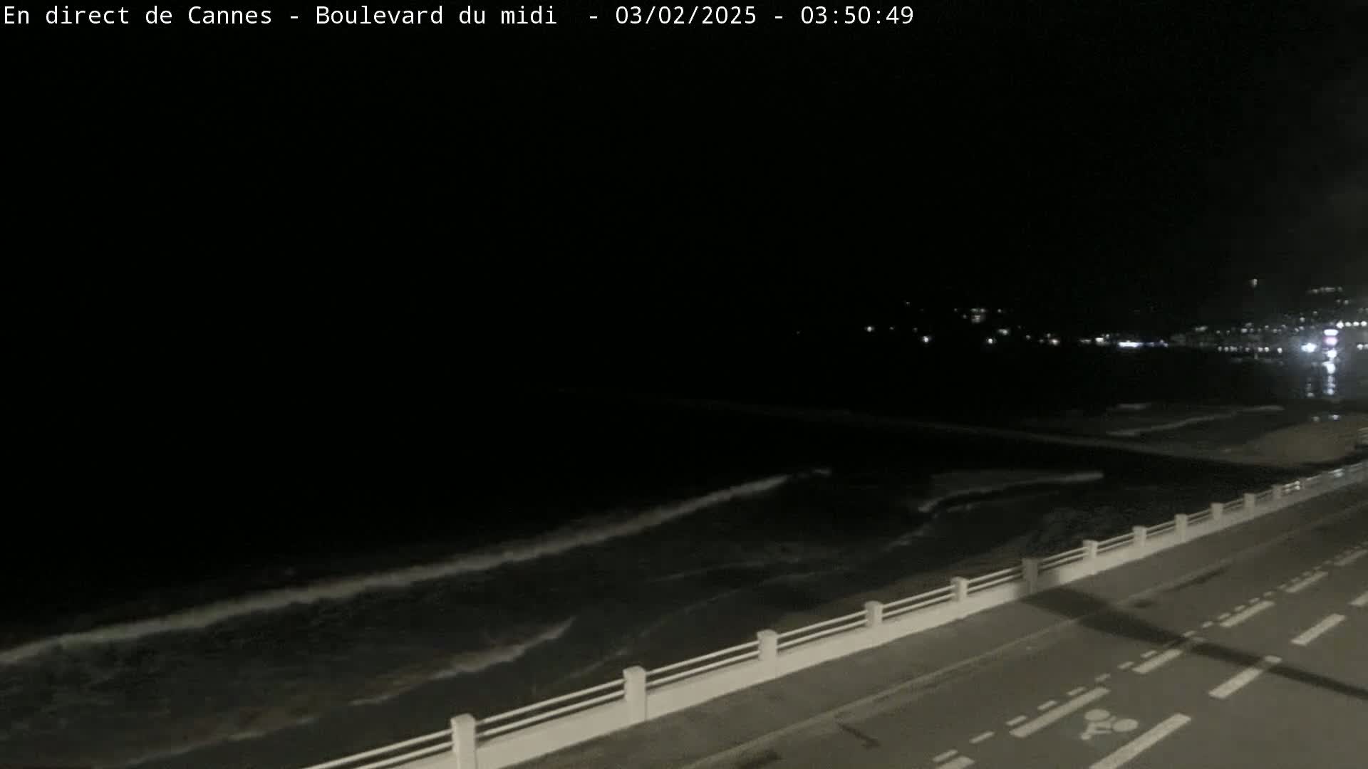 A nighttime, seaside view shows dark water with small waves lapping against a seawall next to a road.