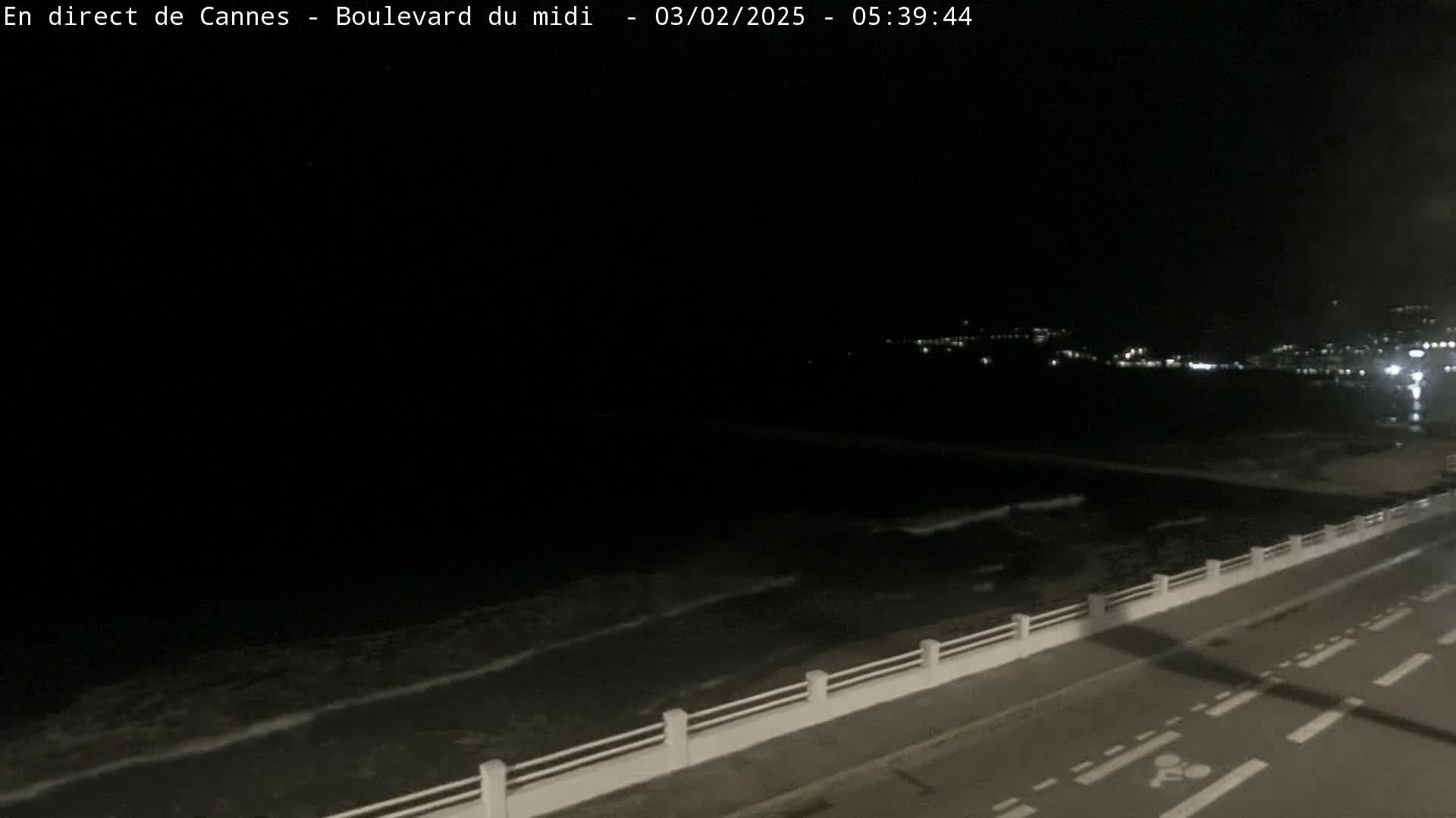 A dark nighttime scene shows a coastline with gently lapping waves, a road with a low white barrier, and distant city lights.