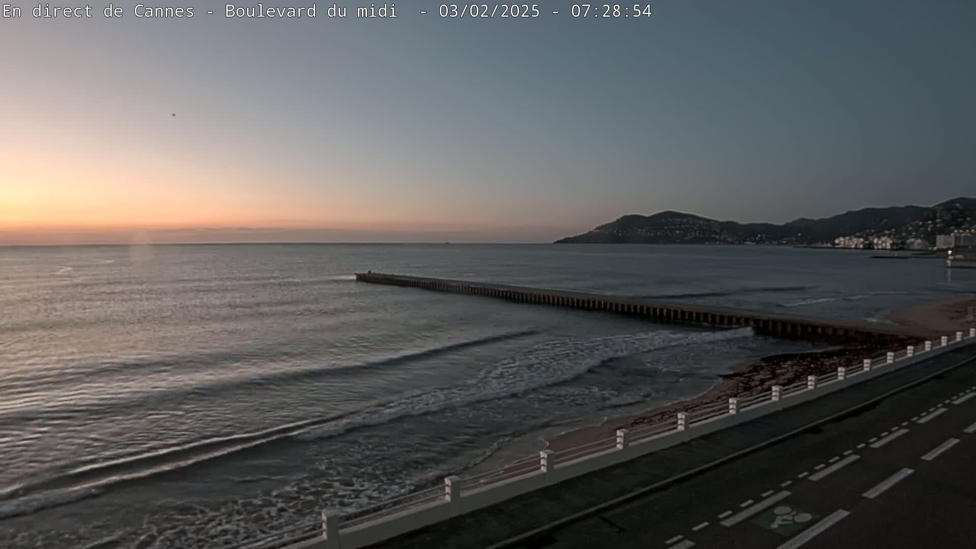 A long pier extends into a calm sea during sunrise, with a coastal road and hills visible in the background.