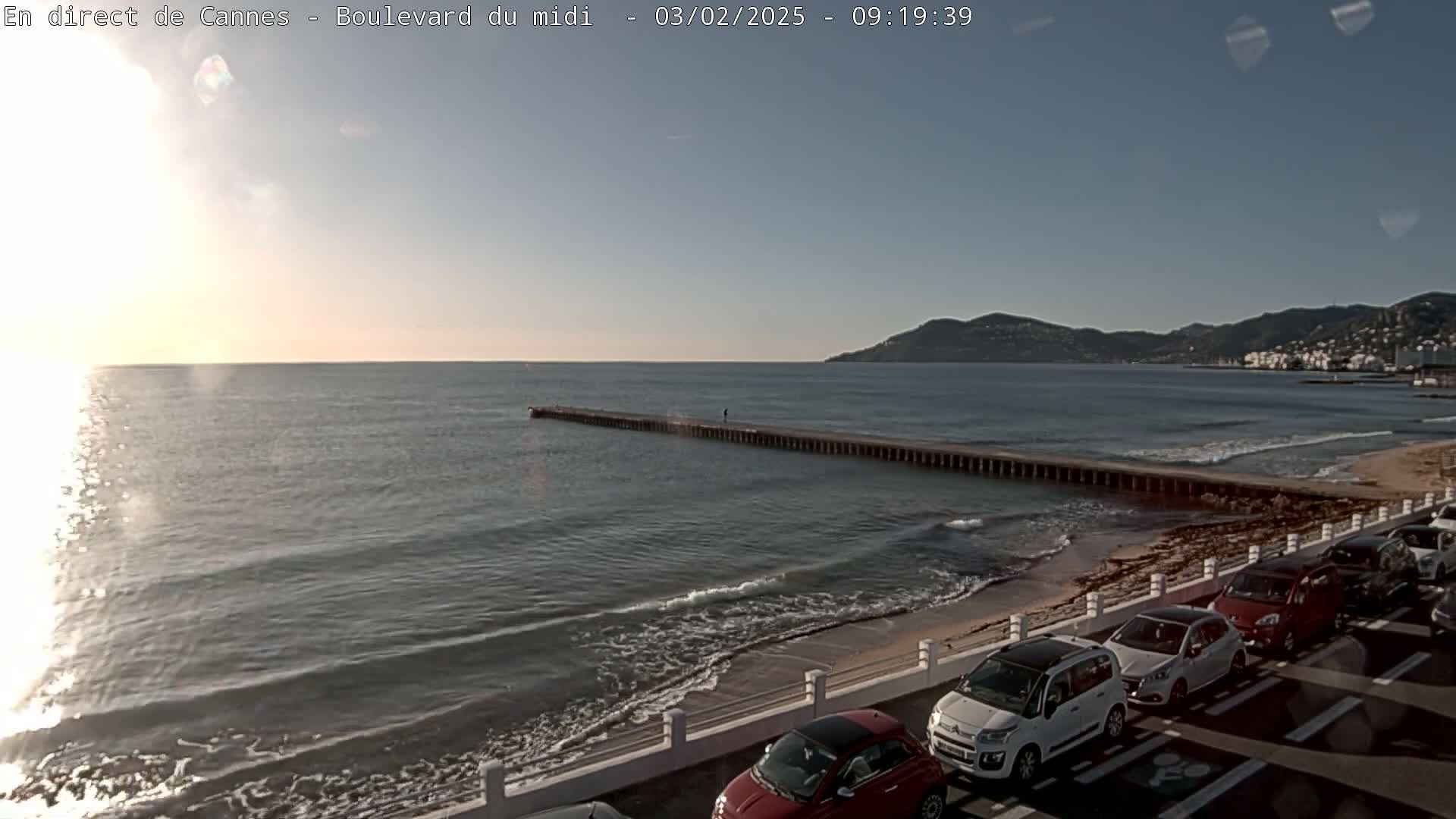A long pier extends into a calm sea under a mostly clear sky, with cars parked along a beach in the foreground.