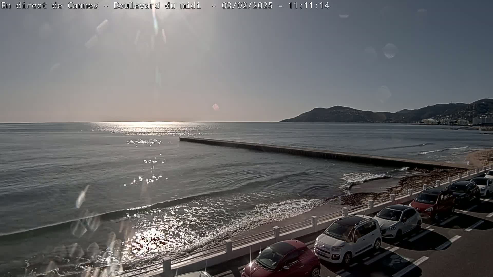A sunny day shows a calm sea with small waves lapping a shore where several cars are parked along a low wall.