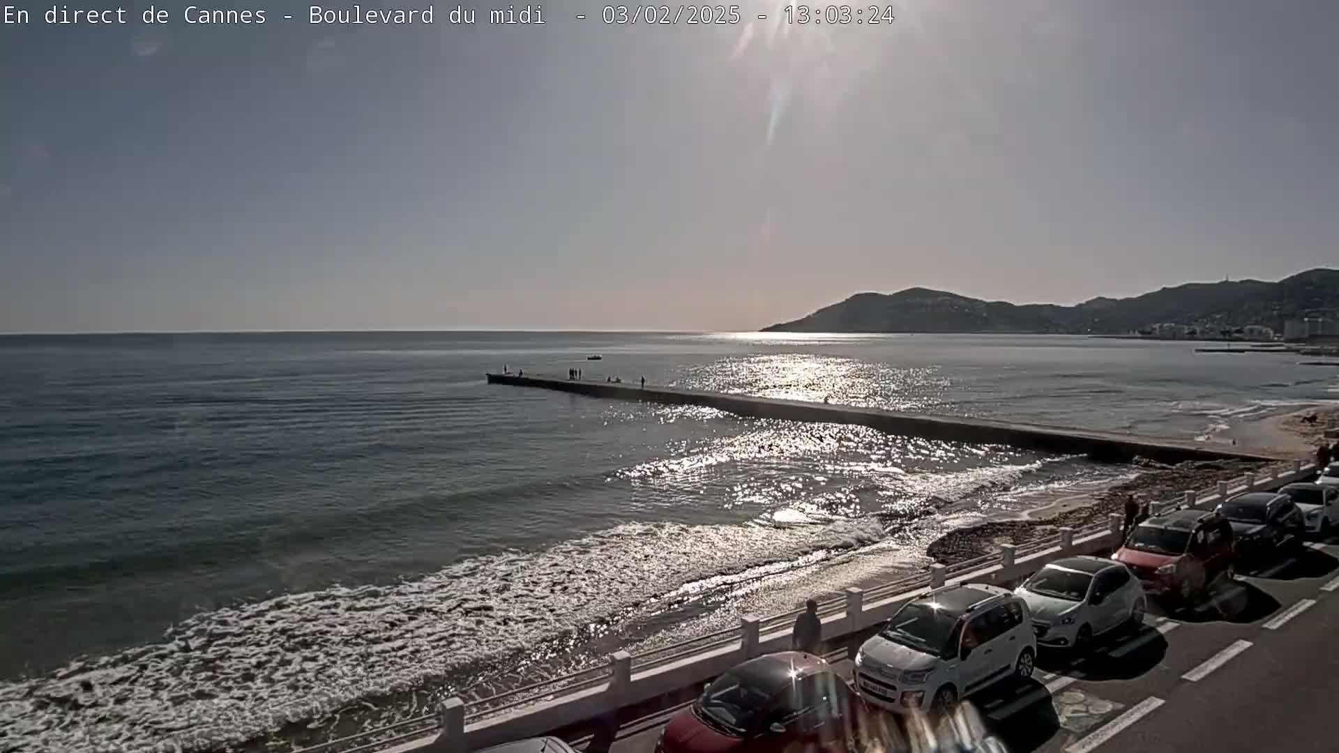 A sunny day at a beach with a long pier extending into the ocean, several cars parked along the roadside, and a few people scattered along the shore and pier.