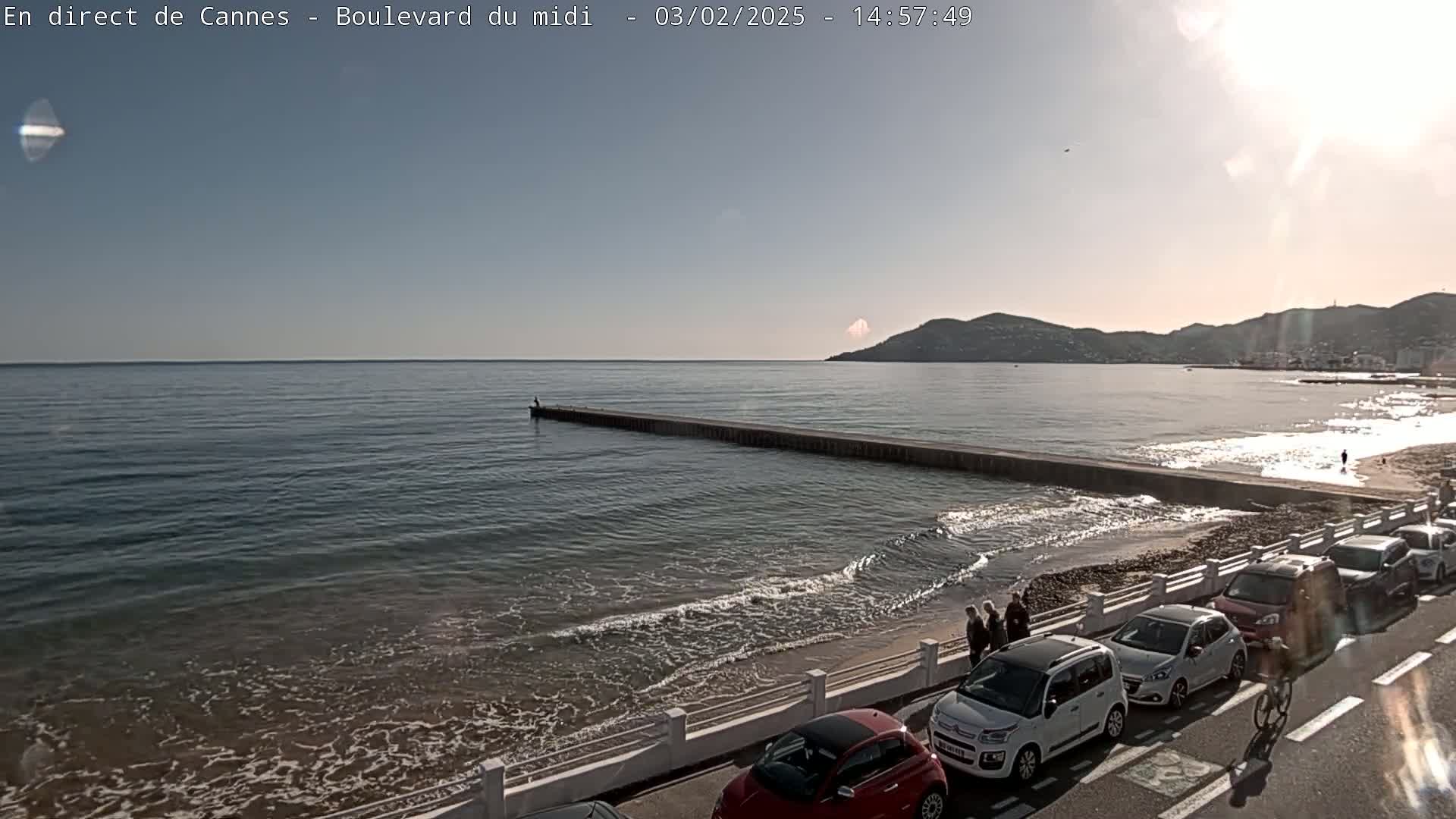 A sunny day at a beach with calm waves, a long jetty extending into the water, several cars parked along the roadside, and a person riding a bicycle.