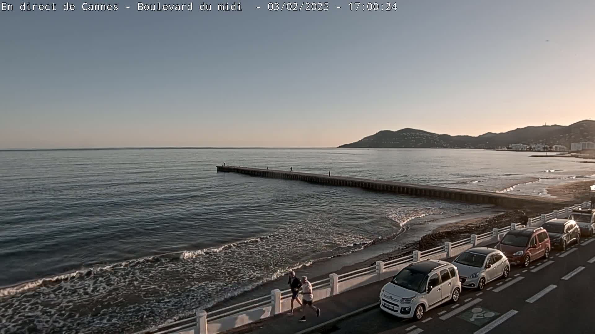 A calm ocean with small waves laps a sandy beach where several cars are parked along a roadway with two people jogging, and a long jetty extends into the water under a clear, pale sky.