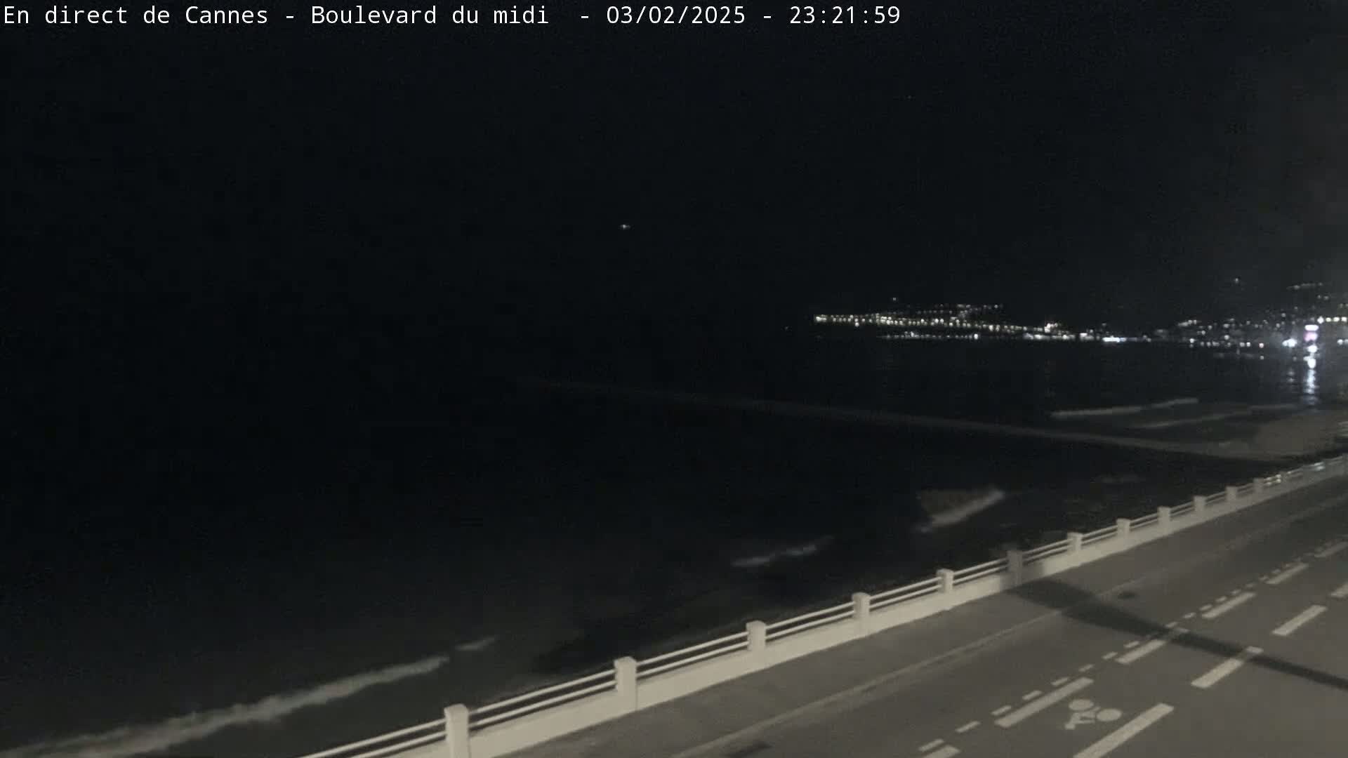 A nighttime view of a dark ocean with gentle waves, a shoreline with a road and a low wall, and a distant city illuminated by lights.