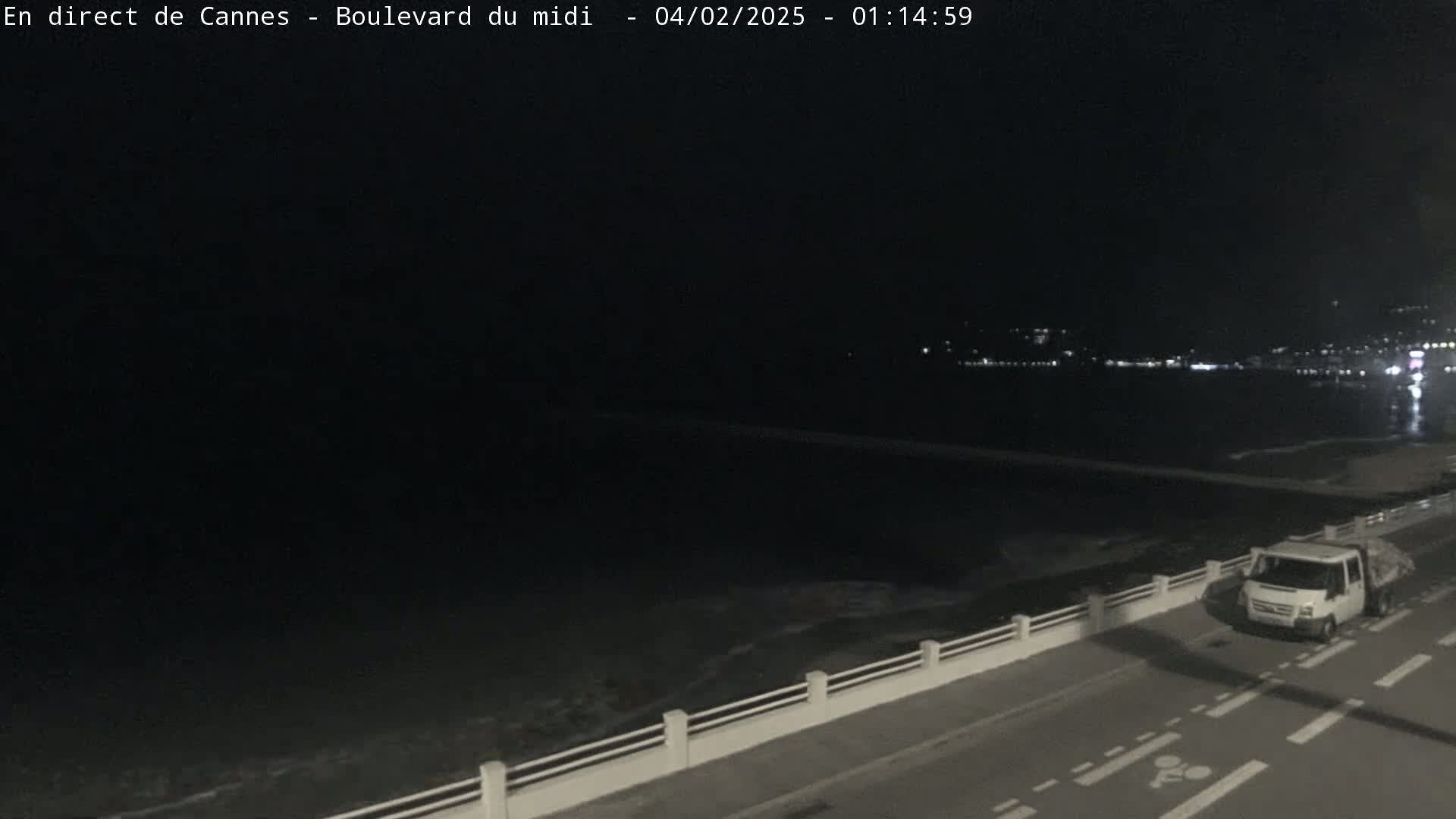 A dark nighttime scene shows a coastal road next to a calm sea, with a single white truck parked on the road.