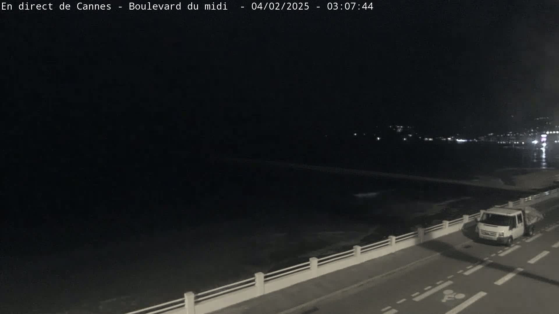A nighttime view of a dark body of water with a coastline showing lights in the distance, a white truck parked on a road next to a low wall, and calm water.