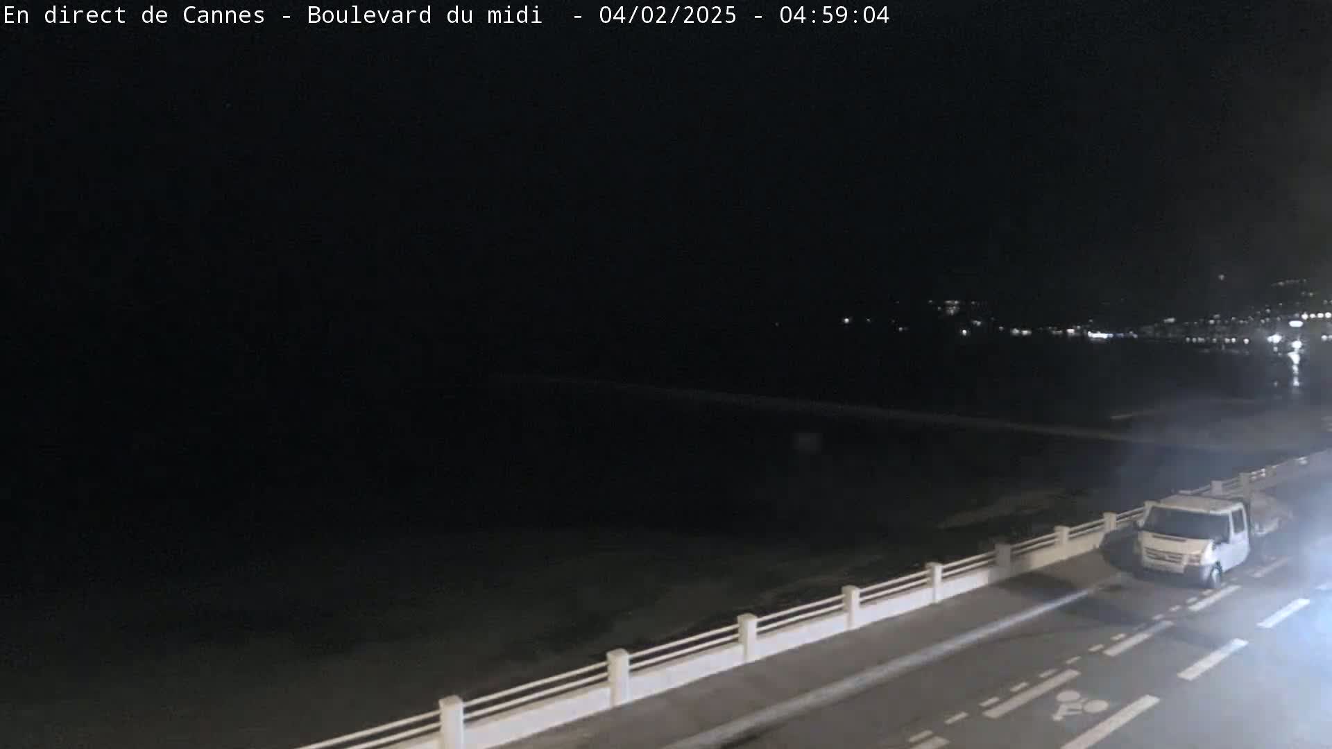 A white van is driving on a dark road at night next to a low white railing overlooking a dark body of water with distant lights in the background.