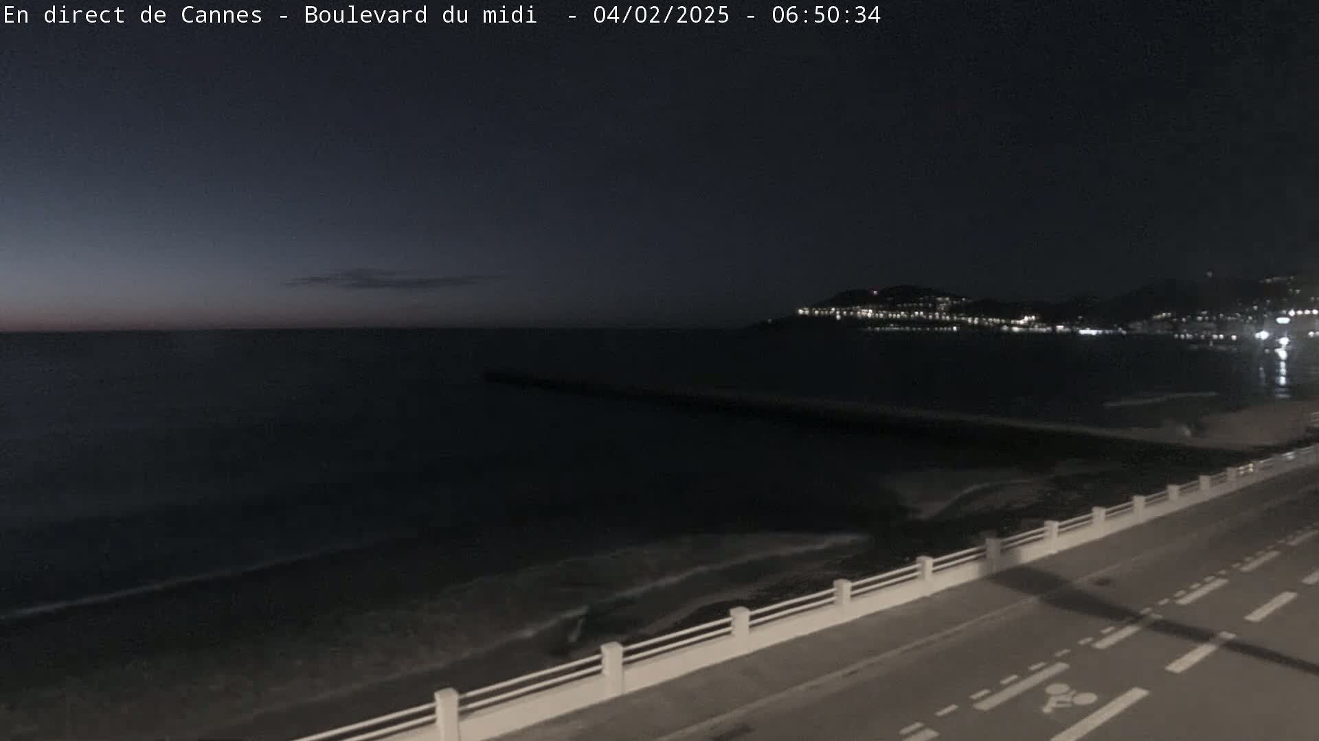 A dark, nighttime seaside scene shows a calm sea with a dimly lit city across the water and a roadway with a low white railing along the shore.