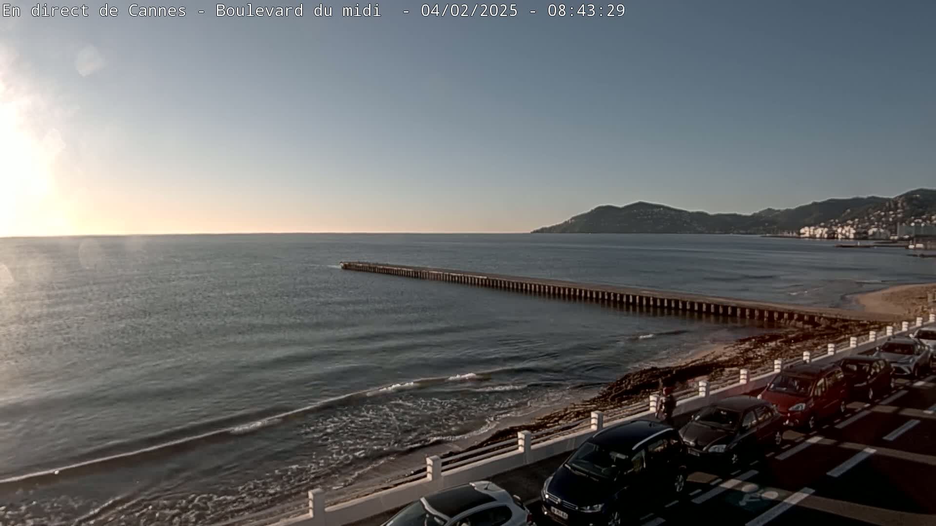 A calm sea with small waves laps a sandy shore where cars are parked next to a low wall, a long pier extends into the water, and hills rise in the distance under a clear sky.