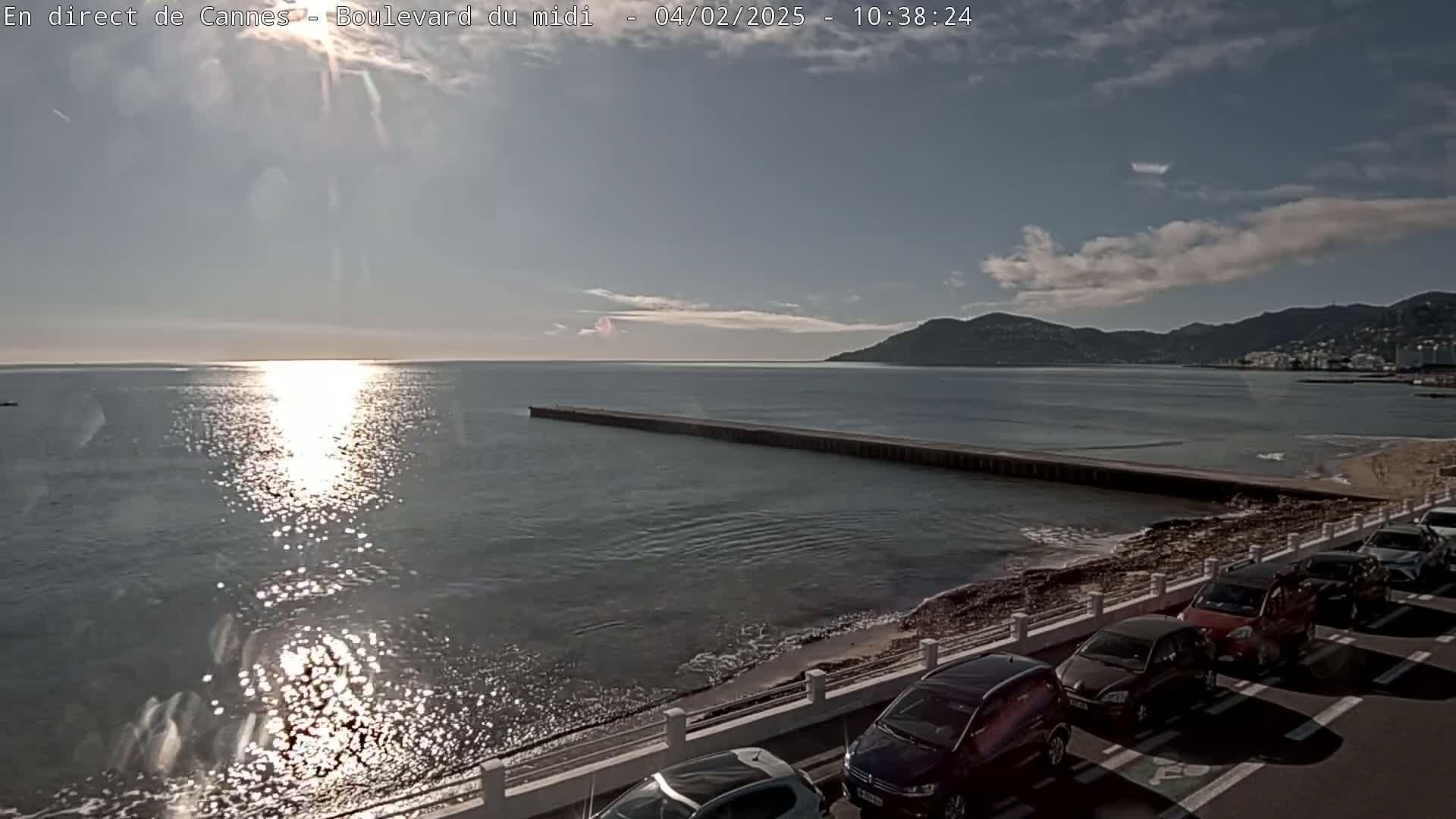 A partly sunny day shows a calm sea reflecting sunlight, a long jetty extending into the water, a shoreline with parked cars, and distant hills under a mostly clear sky.