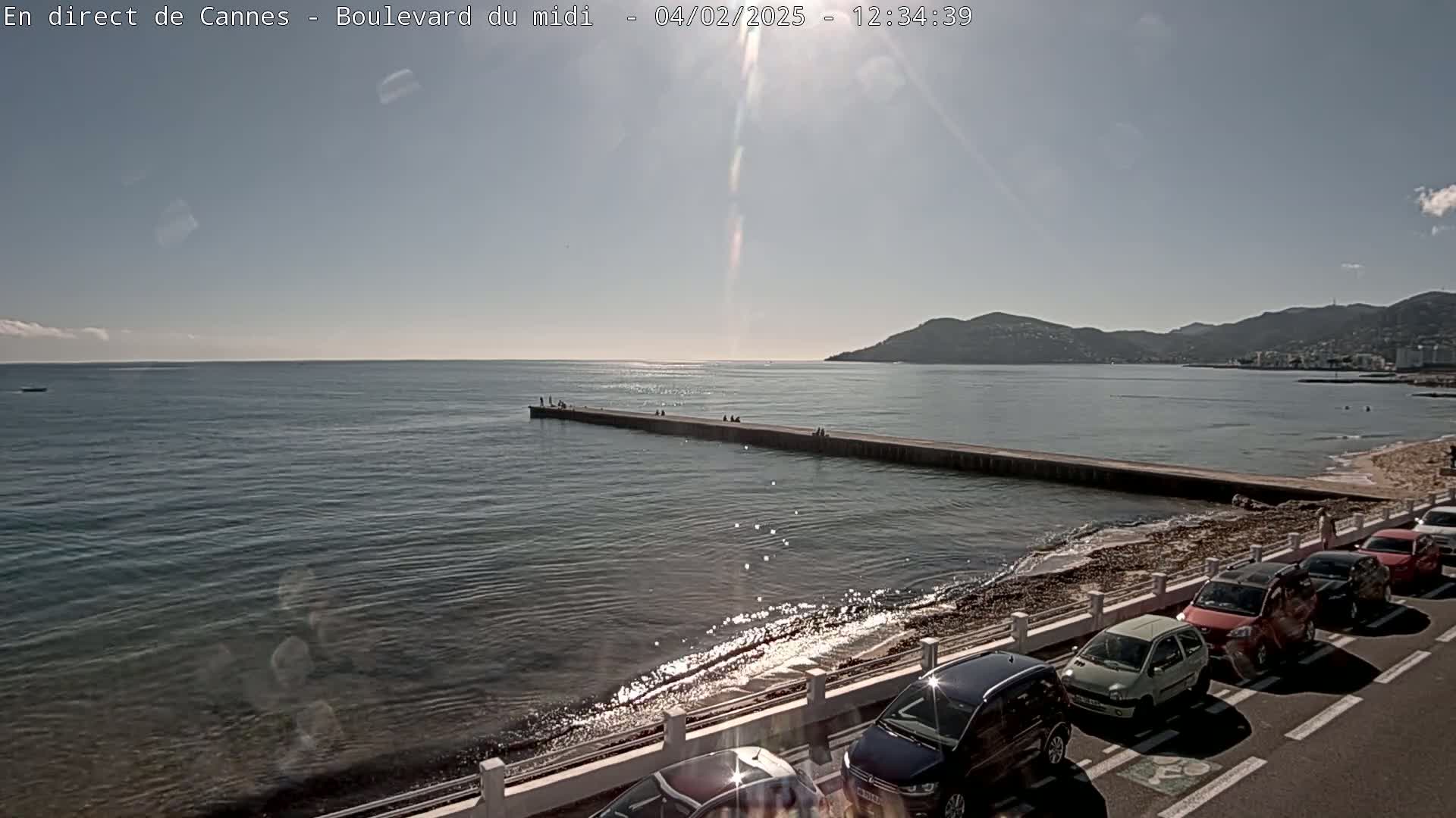 A long pier extends into a calm sea under a bright, sunny sky, with cars parked along a shoreline road and hills visible in the distance.