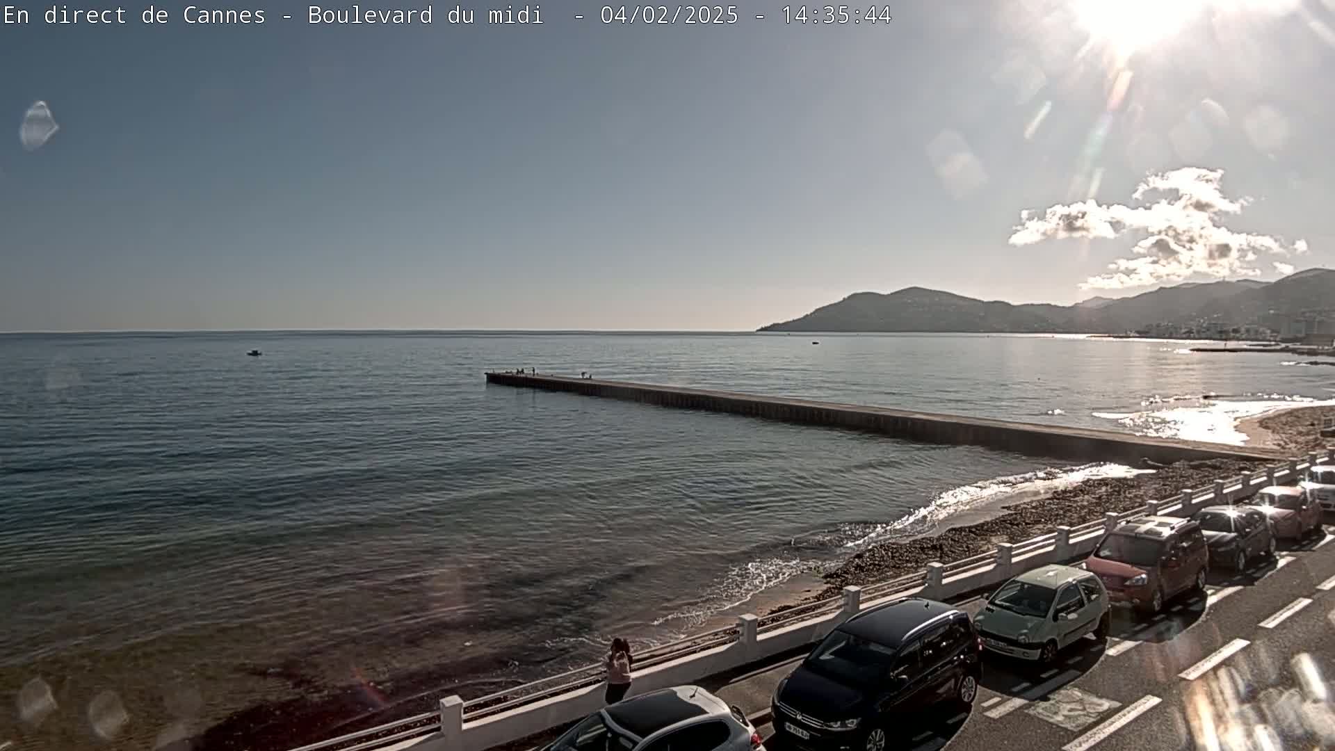 A sunny day at a seaside location shows a long pier extending into a calm sea, with cars parked along a roadway bordering the shore and mountains in the distance.