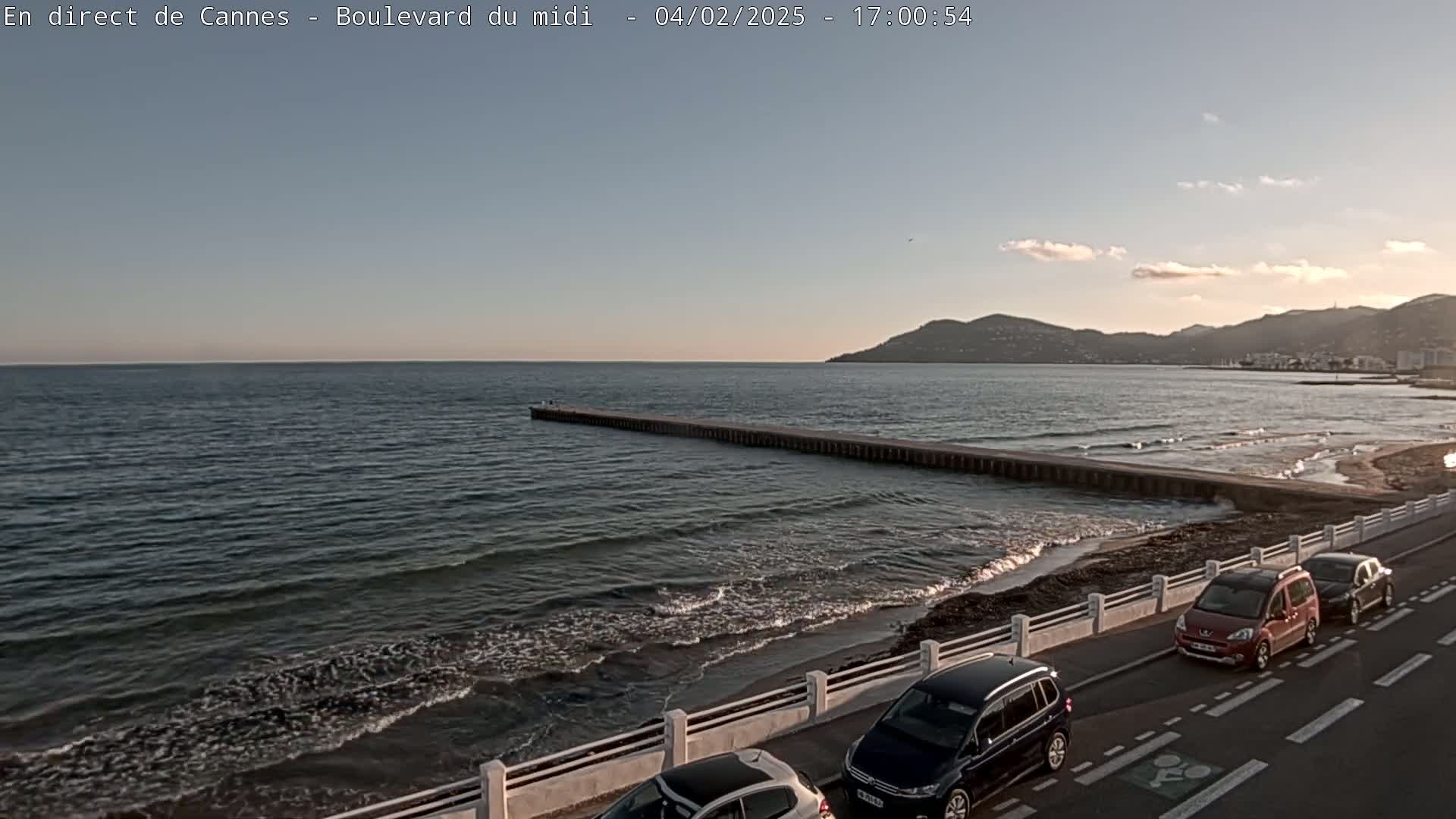 A long pier extends into a calm sea under a partly cloudy sunset sky, with several cars parked along a coastal road.
