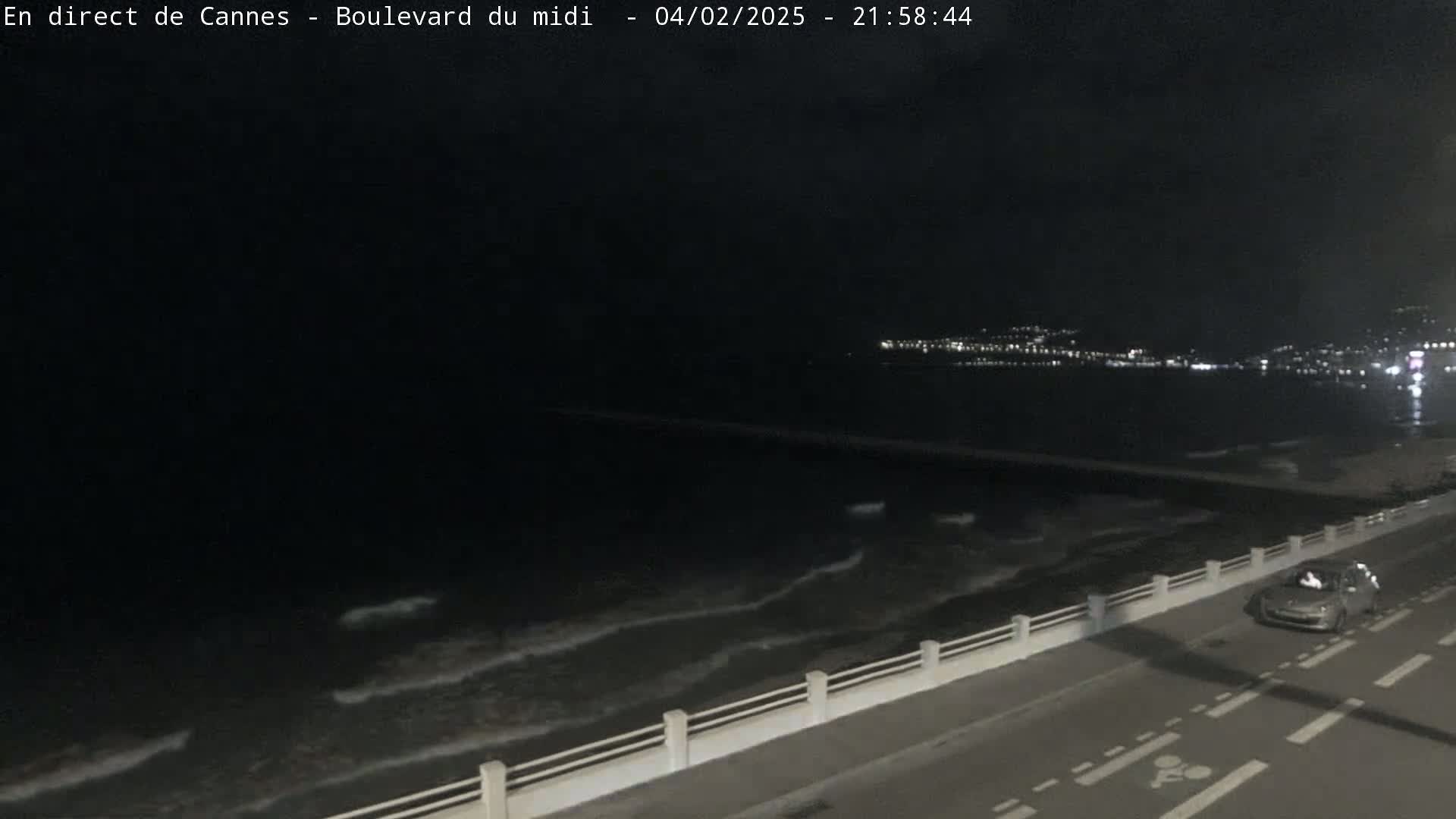 A nighttime view shows a dark ocean with small waves lapping against a seawall next to a road with a single car, and city lights are visible in the distance.