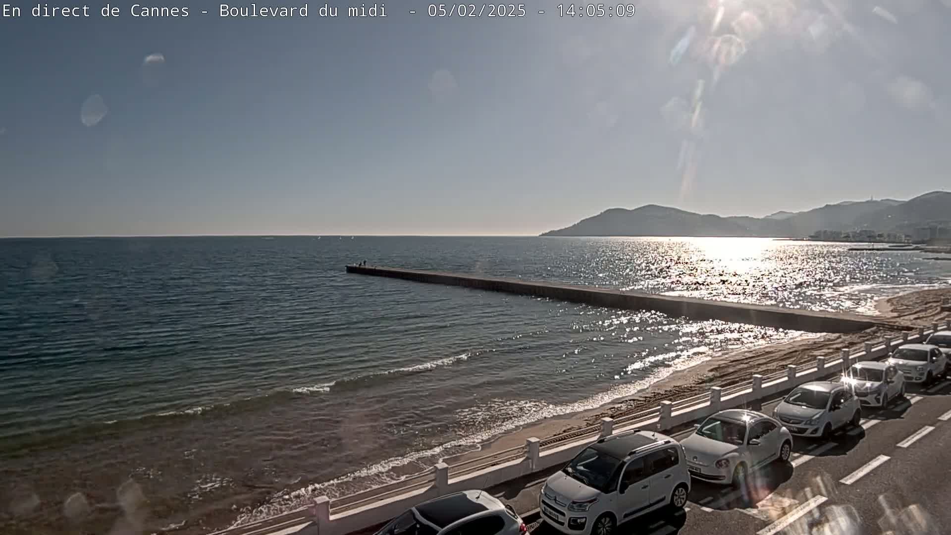 A sunny day at a beach with calm waves, a long jetty extending into the water, and several cars parked along the roadside.