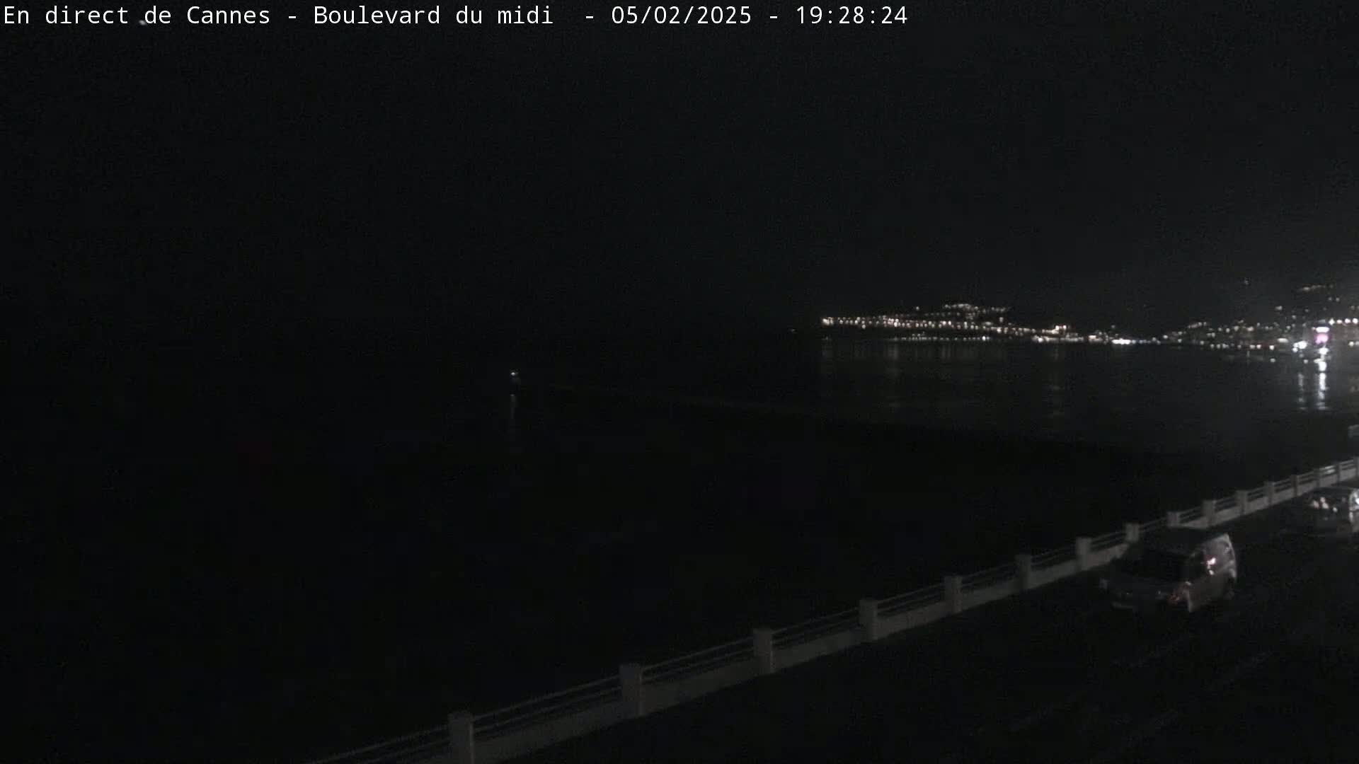 At night, a few vehicles travel along a waterfront road, overlooking a dark body of water and a distant, illuminated city skyline.