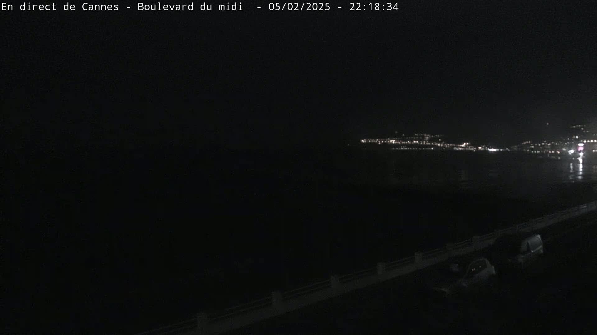 A nighttime, dark view of a coastline with distant city lights across a body of water, a few cars parked nearby.