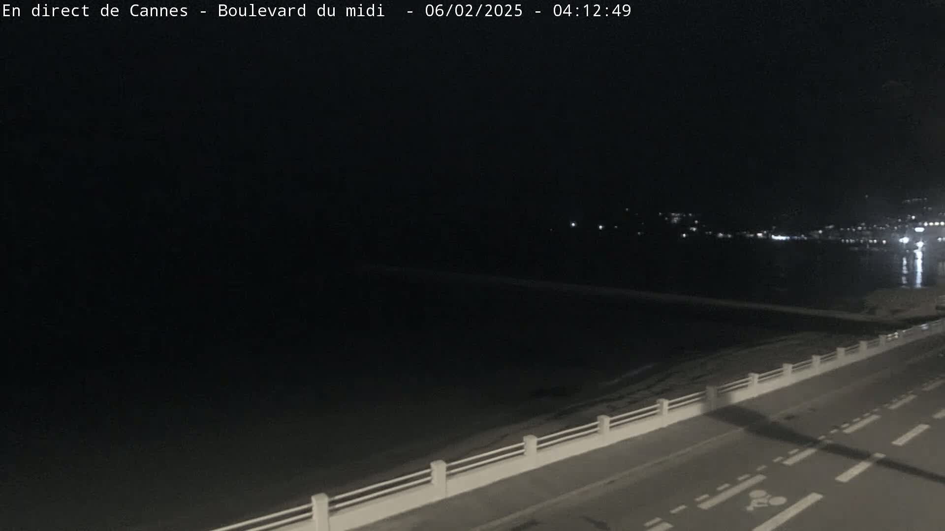 A dark nighttime scene shows a road next to a body of water with a distant shoreline of lights.